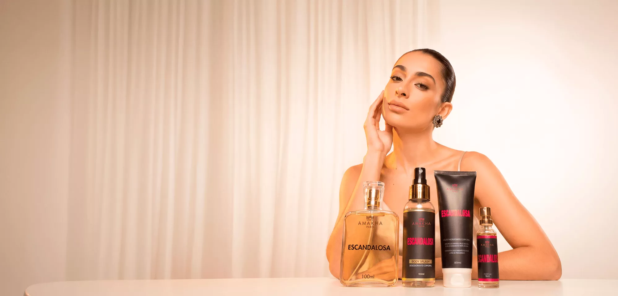 Modelo com a mão no rosto ao lado dos produtos da linha Escandalosa da Amakha Paris: perfume, body splash, hidratante e perfume de bolso