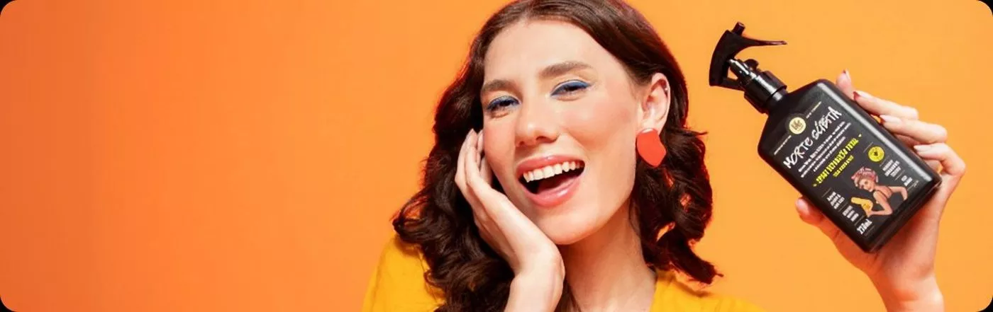Modelo sorrindo segurando o spray Morte Súbita da Lola em fundo laranja